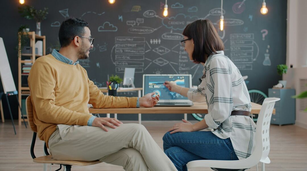Two colleagues at an office desk reviewing charts on an open laptop and talking